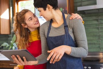 Happy biracial lesbian couple preparing food, using tablet and embracing in kitchen