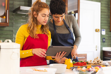 Focused biracial lesbian couple using tablet and preparing vegetables in kitchen