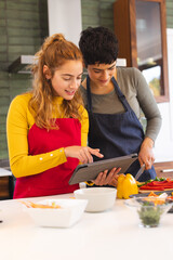 Happy biracial lesbian couple using tablet preparing vegetables in kitchen, copy space