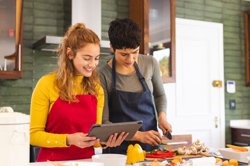 Happy biracial lesbian couple using tablet preparing vegetables in kitchen