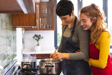 Happy biracial lesbian couple cooking on hob and embracing in kitchen, copy space