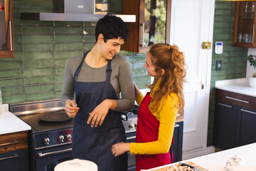 Happy biracial lesbian couple in aprons cooking, talking in kitchen