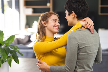 Happy biracial lesbian couple embracing and smiling in kitchen at home - obrazy, fototapety, plakaty