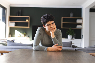 Serious biracial woman sitting at dining table having video call at home, listening