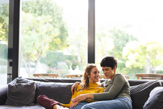 Happy biracial lesbian couple sitting on couch talking in sunny living room, copy space