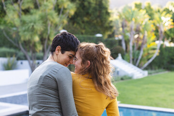 Rear view of happy biracial lesbian couple in garden embracing and touching heads, copy psace