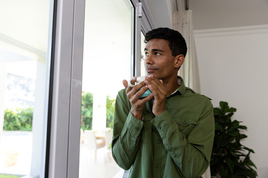 Thoughtful biracial man holding mug looking through window at home