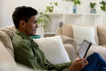 Happy biracial man sitting on sofa using tablet at home