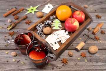 Christmas mulled wine. A pair of glass mugs of mulled wine next to a wooden tray with fruit and Christmas decorations are among various nuts and spices on a gray wooden background.