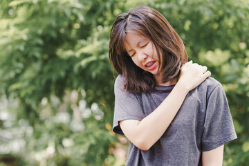 Asian woman holding her shoulder in pain as a result of office syndrome with a natural background. - obrazy, fototapety, plakaty