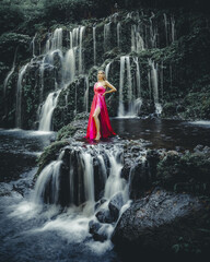 Young Caucasian woman at waterfall in tropical forest. Beautiful slim woman wearing long red dress....