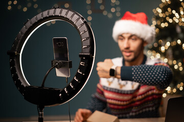 Arab man in Santa hat recording video on phone camera while unpacking box with new smart watch. Male influencer sharing with subscribers his positive feedback about new order. - obrazy, fototapety, plakaty