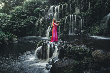 Beautiful Caucasian woman with violin at waterfall in tropical forest. Music and art concept. Female wearing long red dress and playing violin in nature. Slow shutter speed, motion photography.