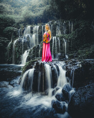 Beautiful Caucasian woman with violin at waterfall in tropical forest. Music and art concept. Female wearing long red dress and holding violin in nature. Slow shutter speed, motion photography.
