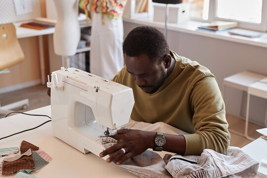 Young African American couturier using electric sewing machine while creating items for new seasonal fashion collection in workshop