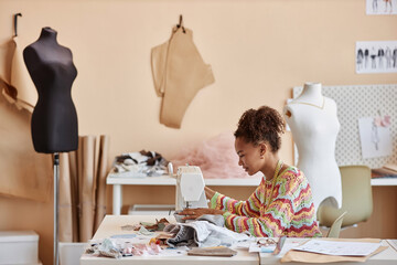 Long shot of young African American female artisan upcycling old clothes in workshop while sitting...