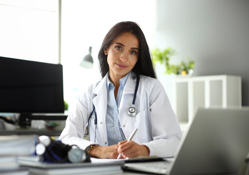 Pretty mature female GP sitting at worktable making notes looking in camera portrait