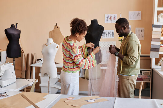 Two young African American couturiers with piece of fatin standing by mannequin while one of them pinning fabric during work