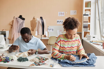 Two young African American stylists creating embroidery on fabric and back of denim jacket while...