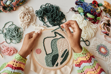 Above view of hands of young female designer making creative embroidery on piece of white canvas...