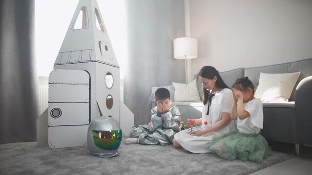 Family day, asian woman with kids play in the living room at home, a boy in an astronaut costume sitting on the floor with her mother and sister, woman tells her children about the solar system.