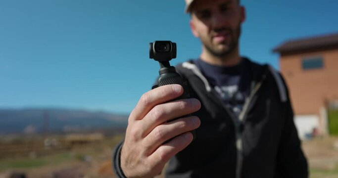 Male Freelancer Holding 3-Axis Stabilized 4K Handheld Camera And Moving Around With Hand. Bokeh Background With Blue Sky 