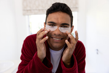 Happy biracial man applying uder-eye patches in bathroom at home