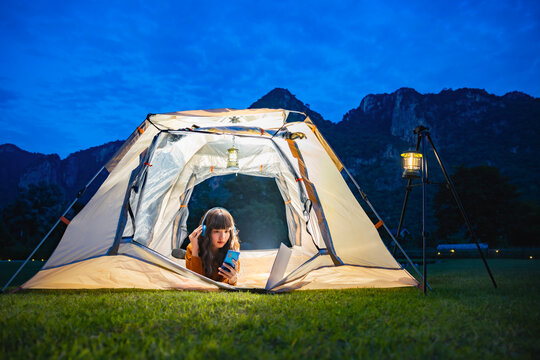 Woman wearing earphones using smartphone to video call with family or friends while lying inside tent. Lamp camping hanging on steel stand. A background of mountain ridge under dark blue sky twilight.
