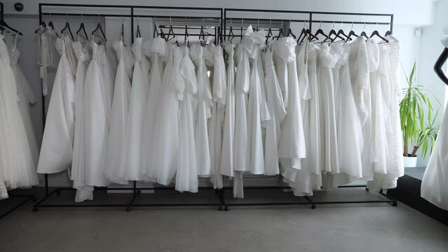 Many different wedding dresses are hanging on hangers in a modern wedding salon. Various wedding dresses. Wedding fashion. Stable movement of the camera backwards. Slow motion.