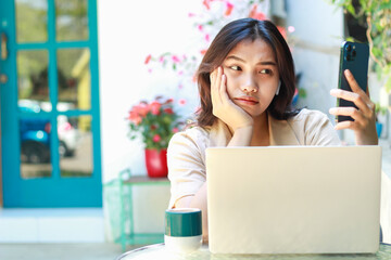 confused asian woman holding smartphone with laptop on table sitting in outdoors cafe working space wearing casual clothes - obrazy, fototapety, plakaty