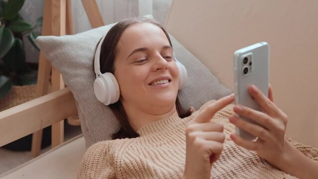 Smiling woman in knitted sweater listening to music with headphones on sofa at home browsing web pages communicating online in social networks.