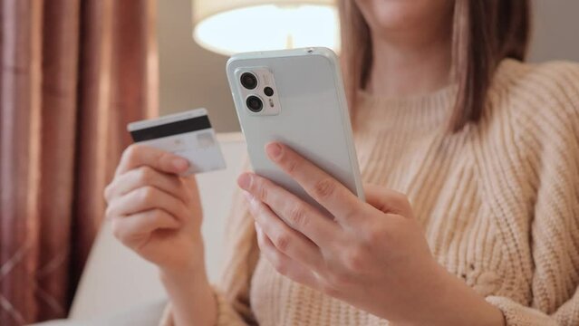 Unrecognizable woman holding smartphone and banking credit card involved in online mobile shopping at home, female shopper purchasing goods or services in internet store.