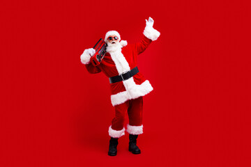 Fototapeta na wymiar Full length photo of positive santa claus in sunglass raising arm up boombox on shoulder at party isolated on red color background