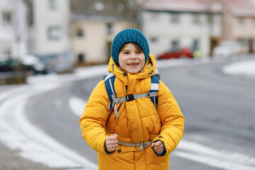 Fototapeta na wymiar Little school kid boy of elementary class walking to school during snowfall. Happy child having fun and playing with first snow. Student with in yellow jacket and backpack in colorful winter clothes.