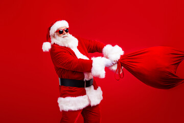 Portrait of good mood funny santa claus in stylish sunglass throwing bag with gifts in empty space isolated on red color background