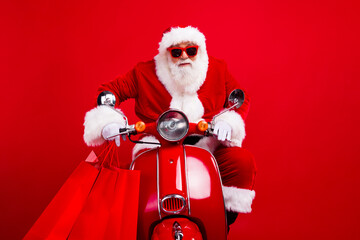 Photo of excited santa claus hold store mall bags drive bike christmas proposition isolated on red color background - obrazy, fototapety, plakaty