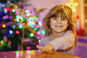 Fototapeta na wymiar Litte toddler girl sitting in pajamas on Christmas morning or eve and with xmas tree and lights on background. Happy healthy child celebrate family traditional holiday, waiting for gifts.