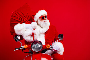 Portrait of positive santa claus with white beard driving scooter hold bag on shoulder look empty space isolated on red color background