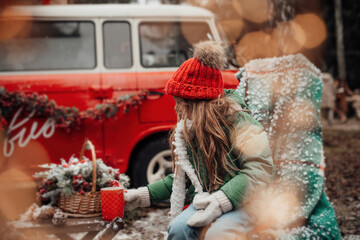 Obraz premium Little girl in red knitted hat drinks hot chocolate from large cup near Xmas bus outdoor