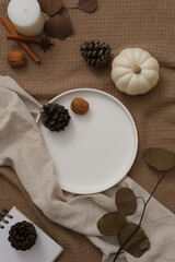 A white ceramic plate is placed in the middle of a brown fabric background. Pumpkins, autumn leaves, pine cones and candles are displayed. Thanksgiving concept. Copy space.