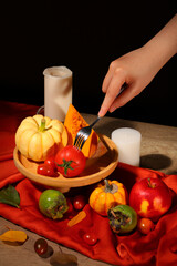 Woman's hand holding a fork stuck into a tomato. Pumpkins and vegetables are displayed on a red cloth on a background with a wooden table. Thanksgiving atmosphere.