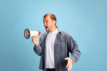 Portrait of angry crazy wild young man shouting to loudspeaker isolated blue studio background. Irritated young guy in casual outfit. Concept of facial expression.