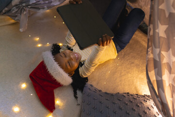 Happy african american girl lying in tappee tent in christmas hat using tablet at home, copy space