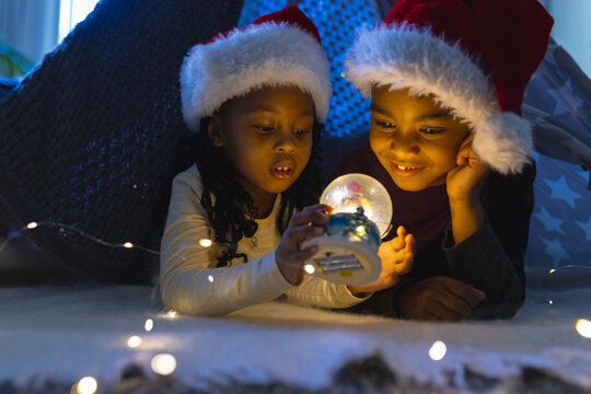 African american boy and girl lying and looking at illuminated christmas snow globe at home - Powered by Adobe