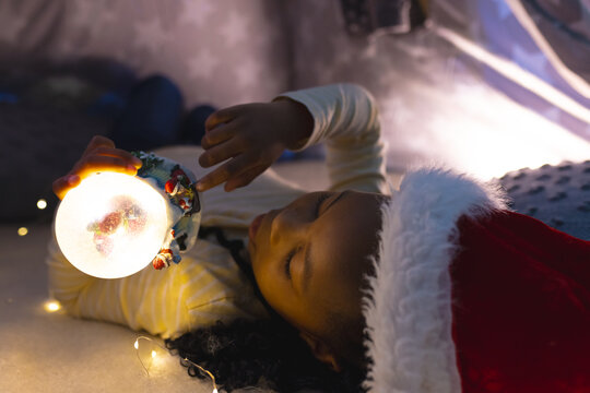African american girl lying, wearing hat and playing with illuminated christmas snow globe at home