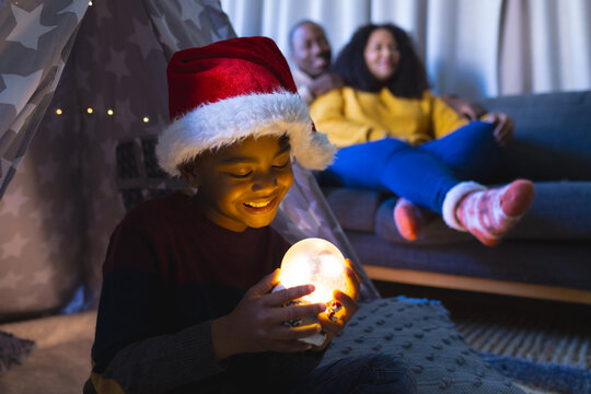 African american boy sitting and playing with illuminated christmas snow globe at home, copy space