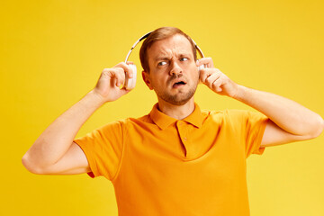 Confused handsome man standing in headphones and can't hear because well music. Guy taking of headphones isolated yellow background.