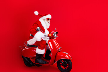 Full length photo of excited santa claus with white beard in sunglass driving scooter look empty space isolated on red color background © deagreez