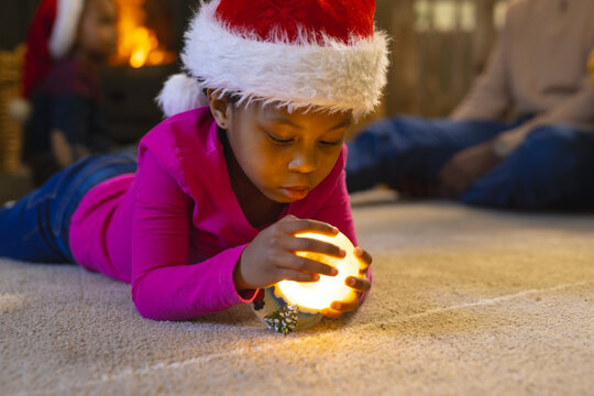 African american girl lying and playing with illuminated christmas snow globe at home, copy space