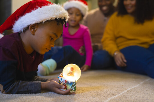 African american boy lying and playing with illuminated christmas snow globe at home, copy space